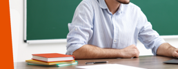 Teacher at a desk on his laptop with a stack of books next to him and a green board behind.
