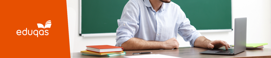 Teacher at a desk on his laptop with a stack of books next to him and a green board behind.