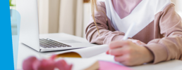 Close up of learner at desk with laptop writing in a notebook.