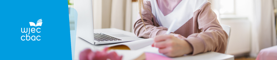 Close up of learner at desk with laptop writing in a notebook.